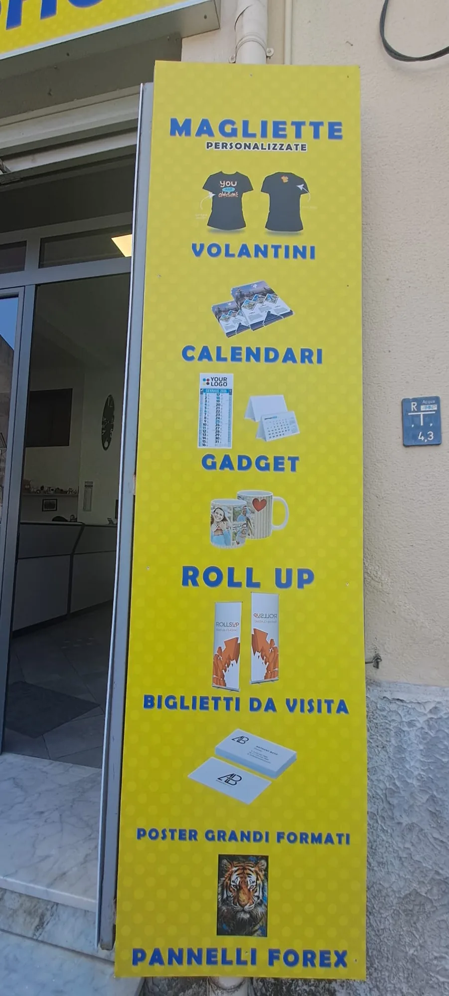 Il totem giallo all'ingresso con l'elenco delle personalizzazioni: magliette, volantini, calendari, gadget, roll up, biglietti da visita, poster grandi formati e pannelli forex