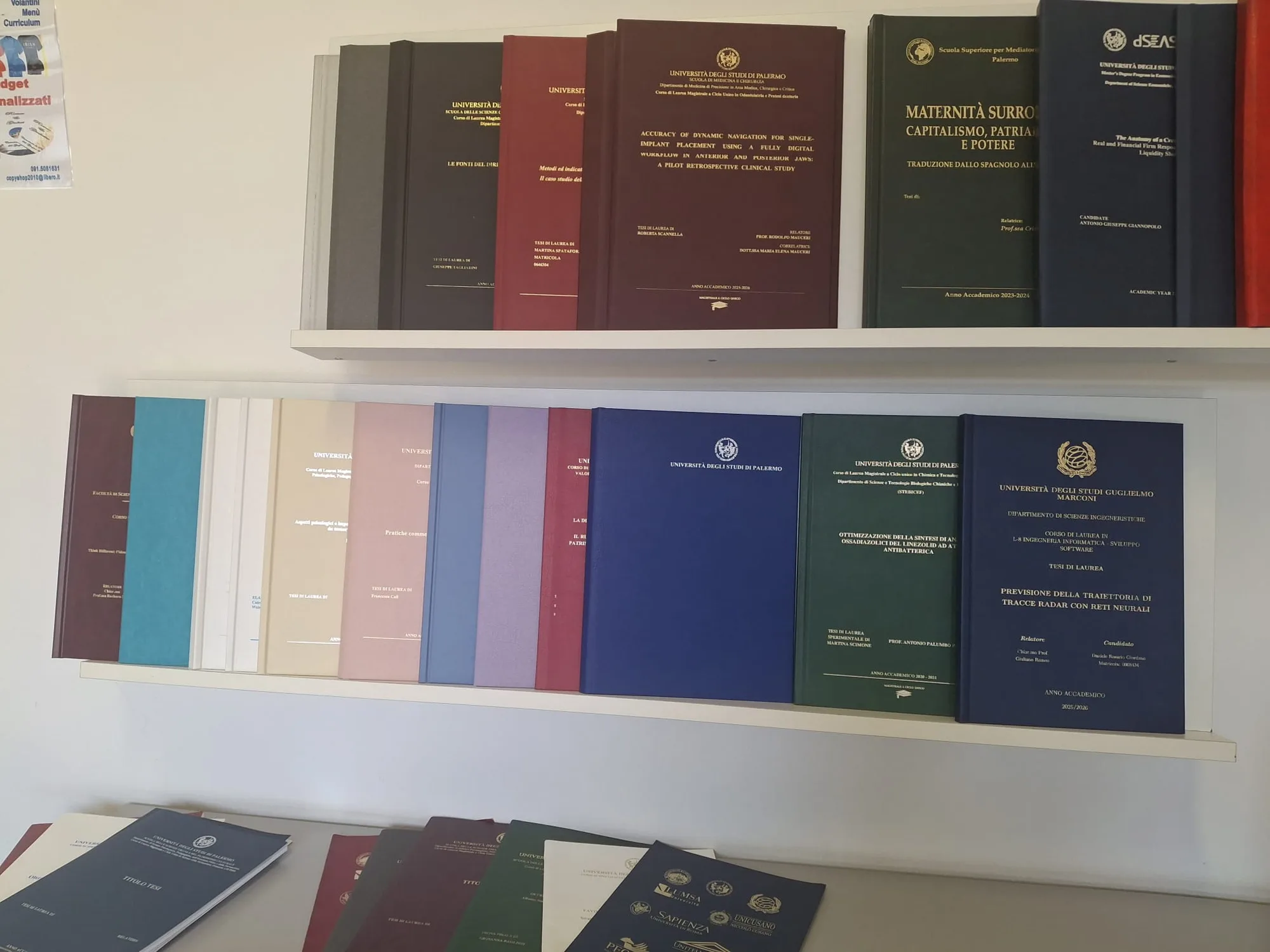 Due ripiani di tesi di laurea rilegate in pelle: bordeaux, blu, verde, grigio, con titolo e ateneo impressi in oro e argento