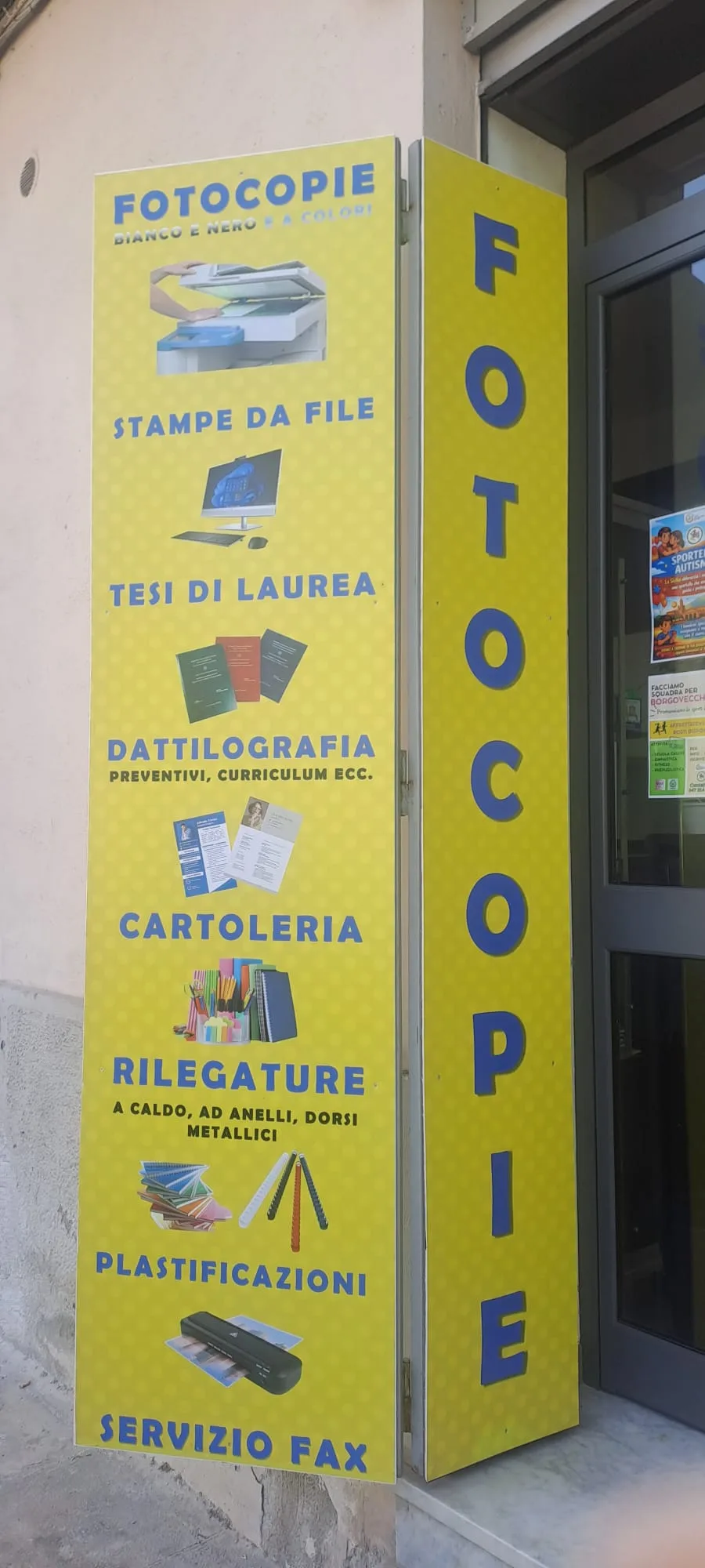 Il totem giallo davanti alla porta con l'elenco dei servizi: fotocopie, stampe da file, tesi di laurea, dattilografia, cartoleria, rilegature, plastificazioni, servizio fax